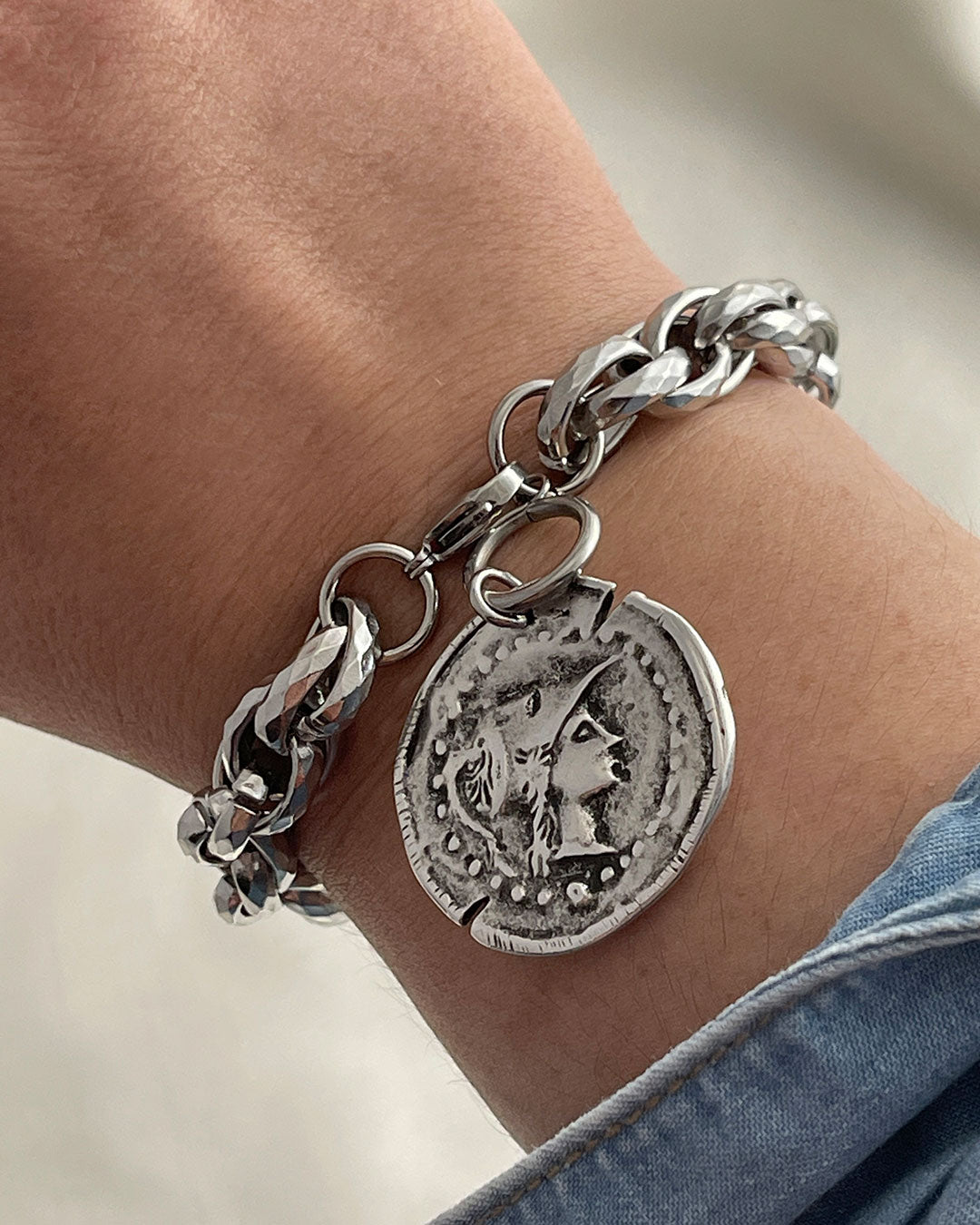 Chunky silver bracelet with an Athena coin charm on a wrist, against a neutral background