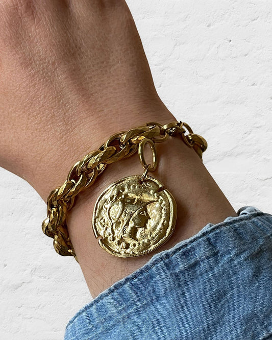 A gold chain bracelet with a large, round, gold coin charm. The coin has a raised design of Athena's goddess face.