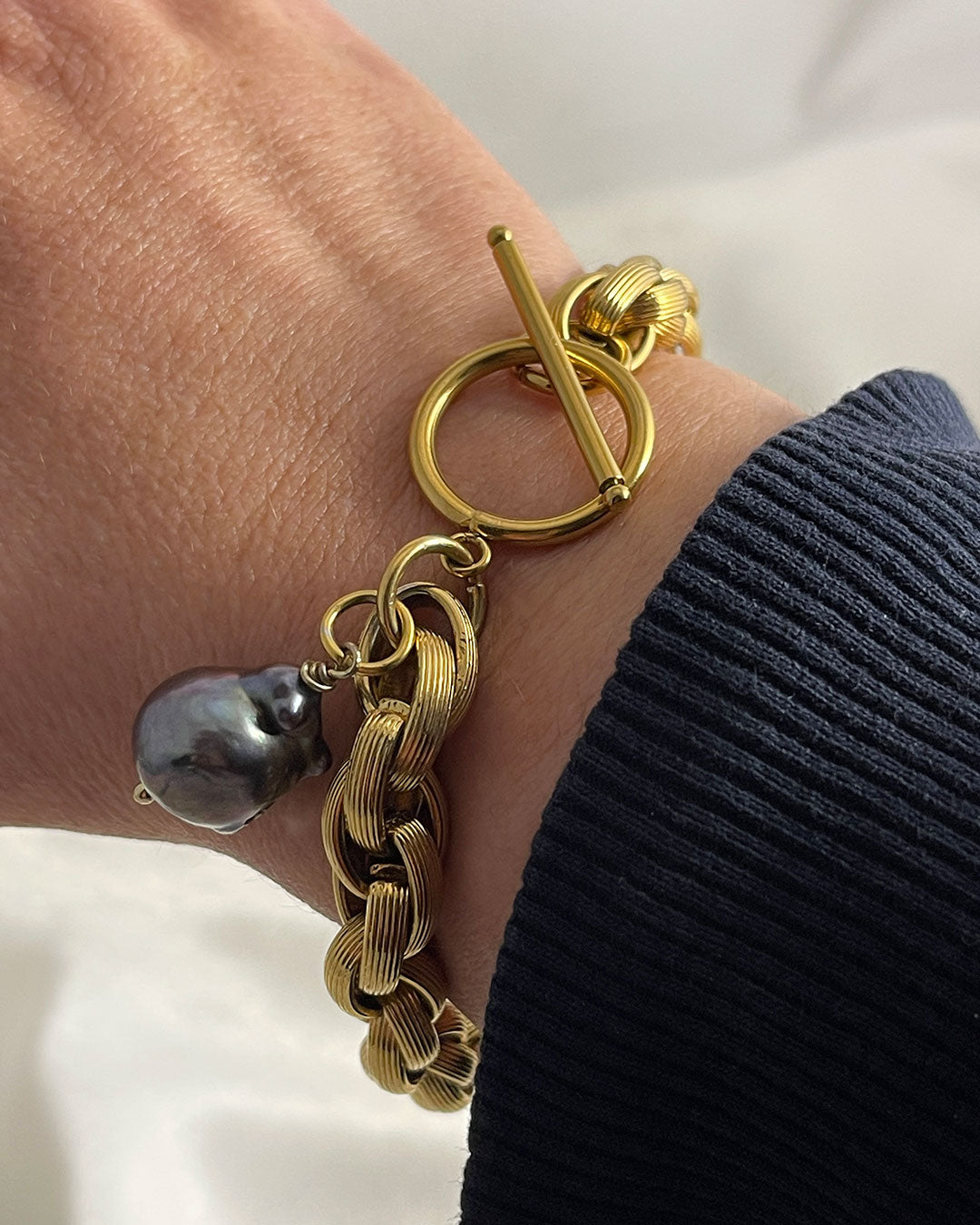 Model wearing a gold toggle bracelet featuring a braided chain design and a dark gray baroque pearl accent. The bracelet has a substantial, chunky link style and a secure clasp. The pearl is a focal point, adding a touch of elegance to the bold gold chain. This statement piece is perfect for adding a touch of luxury to any outfit.