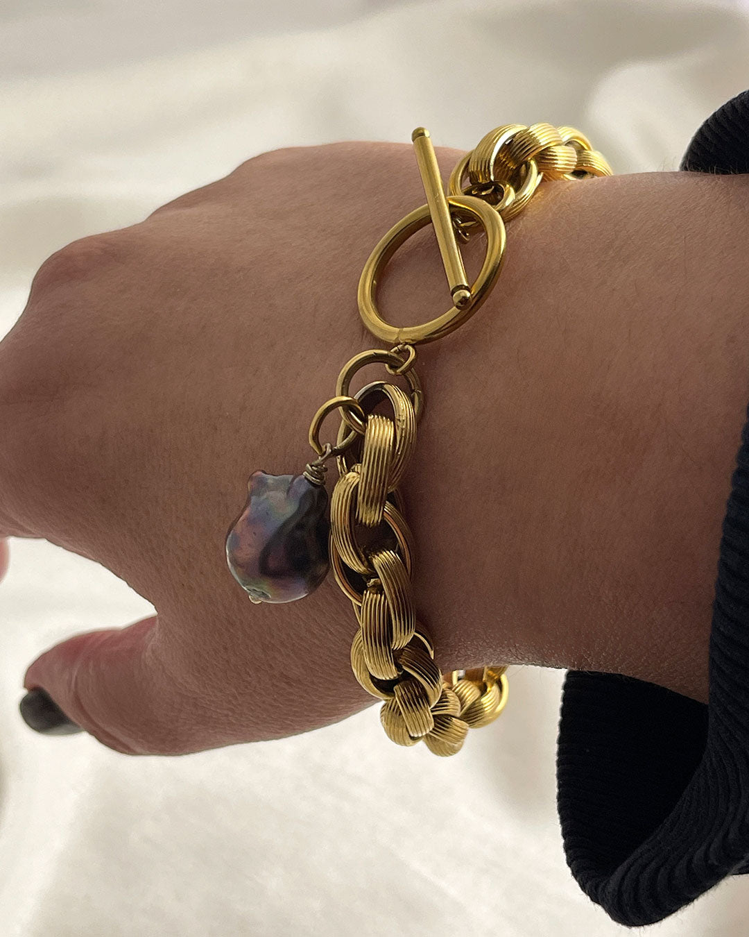 Model wearing a gold toggle bracelet featuring a braided chain design and a dark gray baroque pearl accent. The bracelet has a substantial, chunky link style and a secure clasp. The pearl is a focal point, adding a touch of elegance to the bold gold chain. This statement piece is perfect for adding a touch of luxury to any outfit.