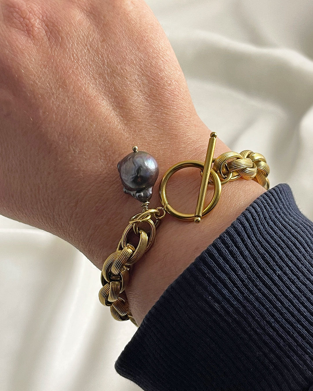 Model wearing a gold toggle bracelet featuring a braided chain design and a dark gray baroque pearl accent. The bracelet has a substantial, chunky link style and a secure clasp. The pearl is a focal point, adding a touch of elegance to the bold gold chain. This statement piece is perfect for adding a touch of luxury to any outfit.