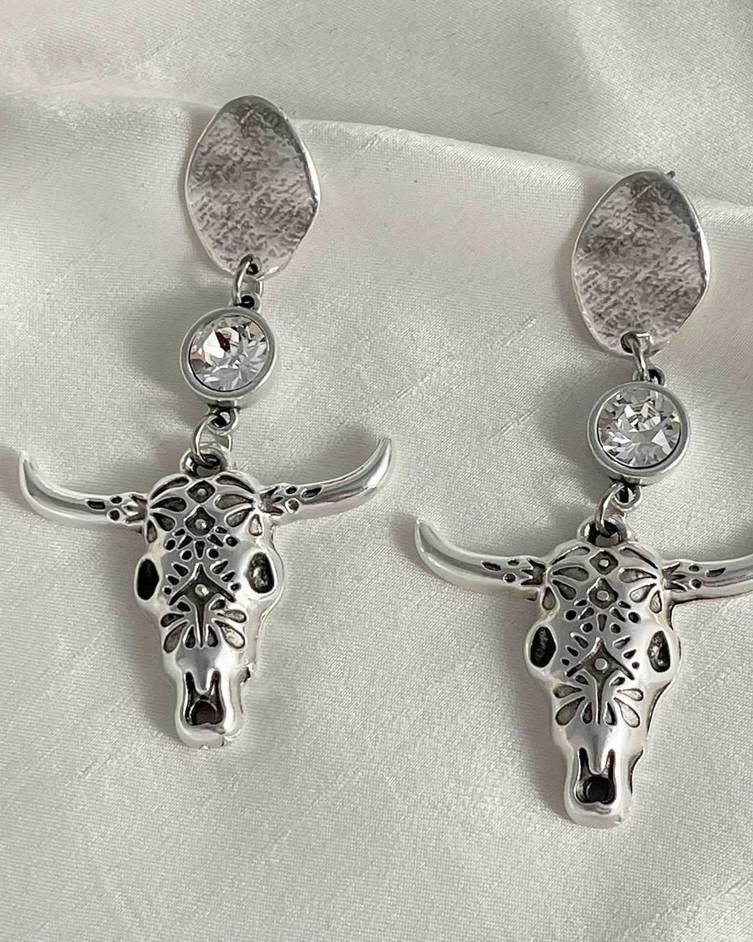 A pair of silver earrings featuring longhorn bull skull large charms. Each pendant is detailed with an embossed design and accented with a crystal clear Swarovski. The earrings hang from silver oval-shaped studs. These Western-style earrings are perfect for adding a touch of bohemian flair to any outfit.