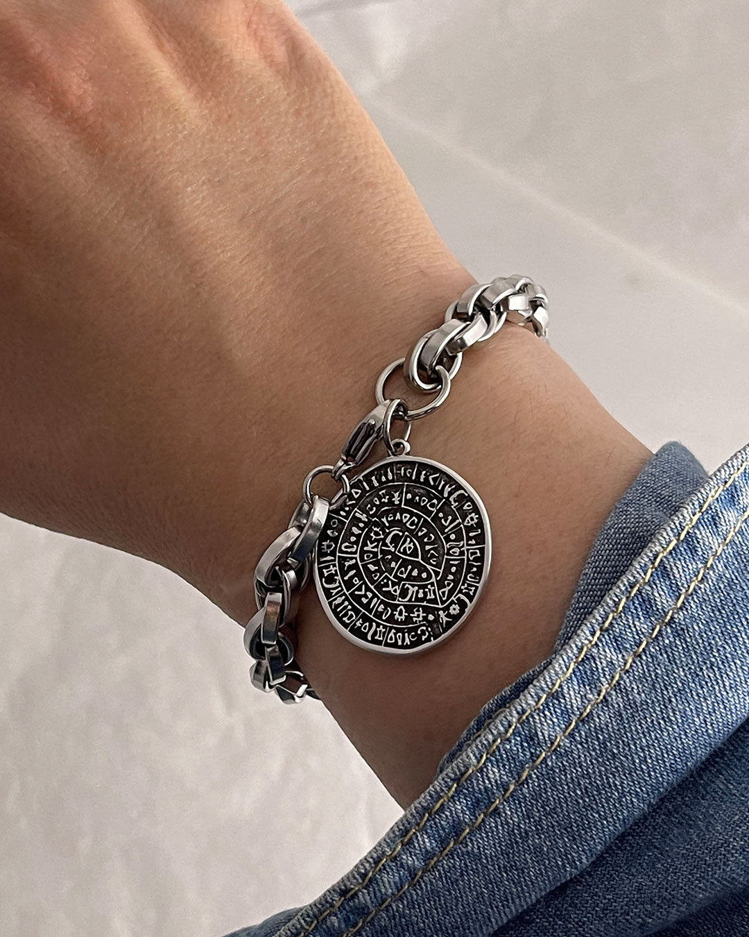 Silver bracelet on the wrist with a round Phaistos disc, against a neutral background