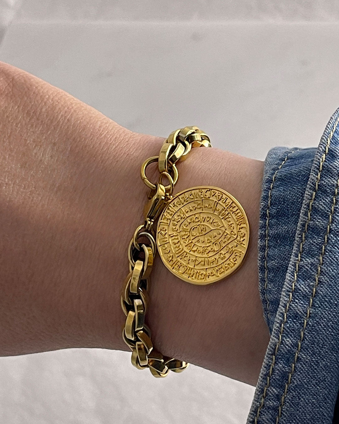Gold bracelet on the wrist with a round Phaistos disc, against a neutral background
