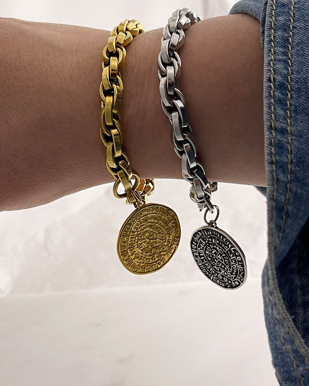 Two bracelets on a wrist, one gold and one silver, with round charms featuring the Phaistos disc. The bracelet has a chunky braided chain design.