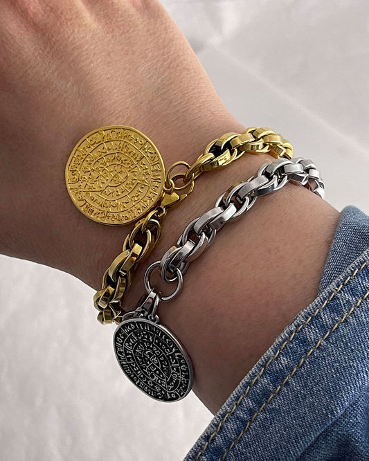 Two bracelets on a wrist, one gold and one silver, with round charms featuring the Phaistos disc. The bracelet has a chunky braided chain design.