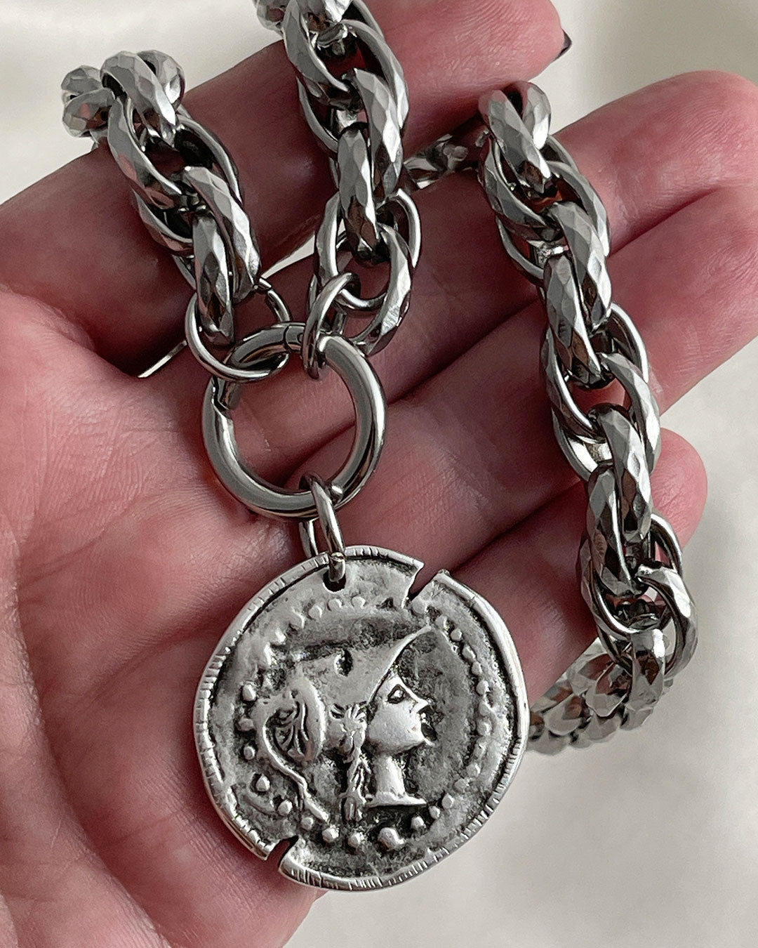 Close-up details from a silver-tone chunky chain necklace with a large coin pendant. The coin features an embossed profile of a woman's face. The necklace is a bold statement piece with a substantial chain and a detailed pendant. The chain is thick and rope-like, and the pendant is substantial and eye-catching.