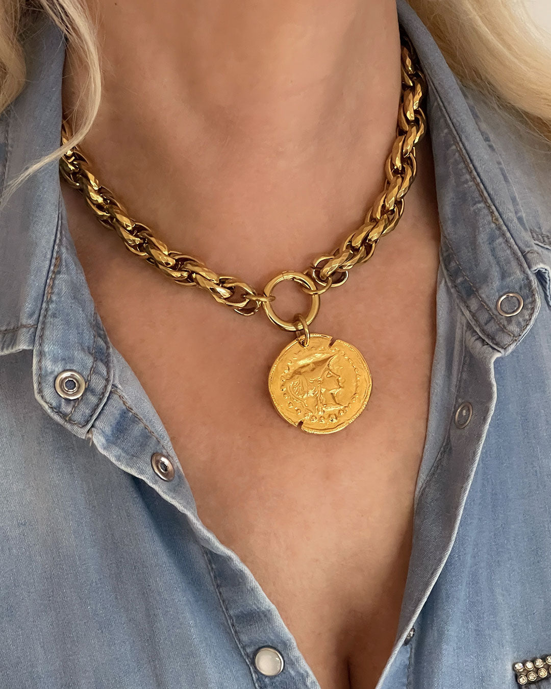 Model wearing a gold tone chunky chain necklace with a large coin pendant. The coin features an embossed profile of a woman's face. The necklace is a bold statement piece with a substantial chain and a detailed pendant. The chain is thick and rope-like, and the pendant is substantial and eye-catching.
