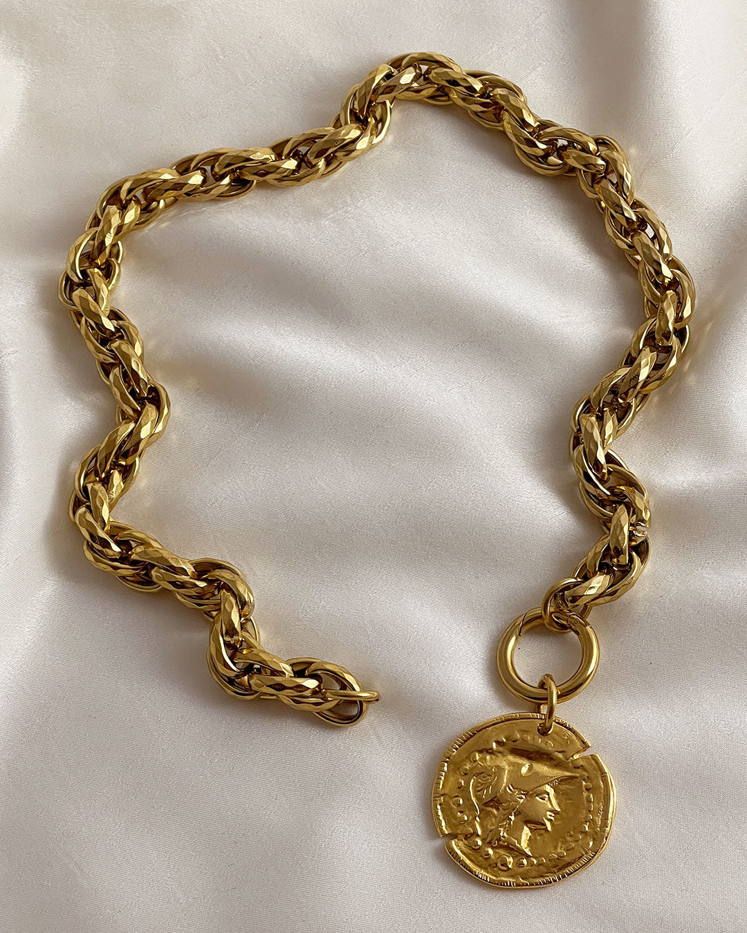 Gold tone chunky chain necklace with a large coin pendant on a beige silk fabric. The coin features an embossed profile of a woman's face. The necklace is a bold statement piece with a substantial chain and a detailed pendant. The chain is thick and rope-like, and the pendant is substantial and eye-catching.