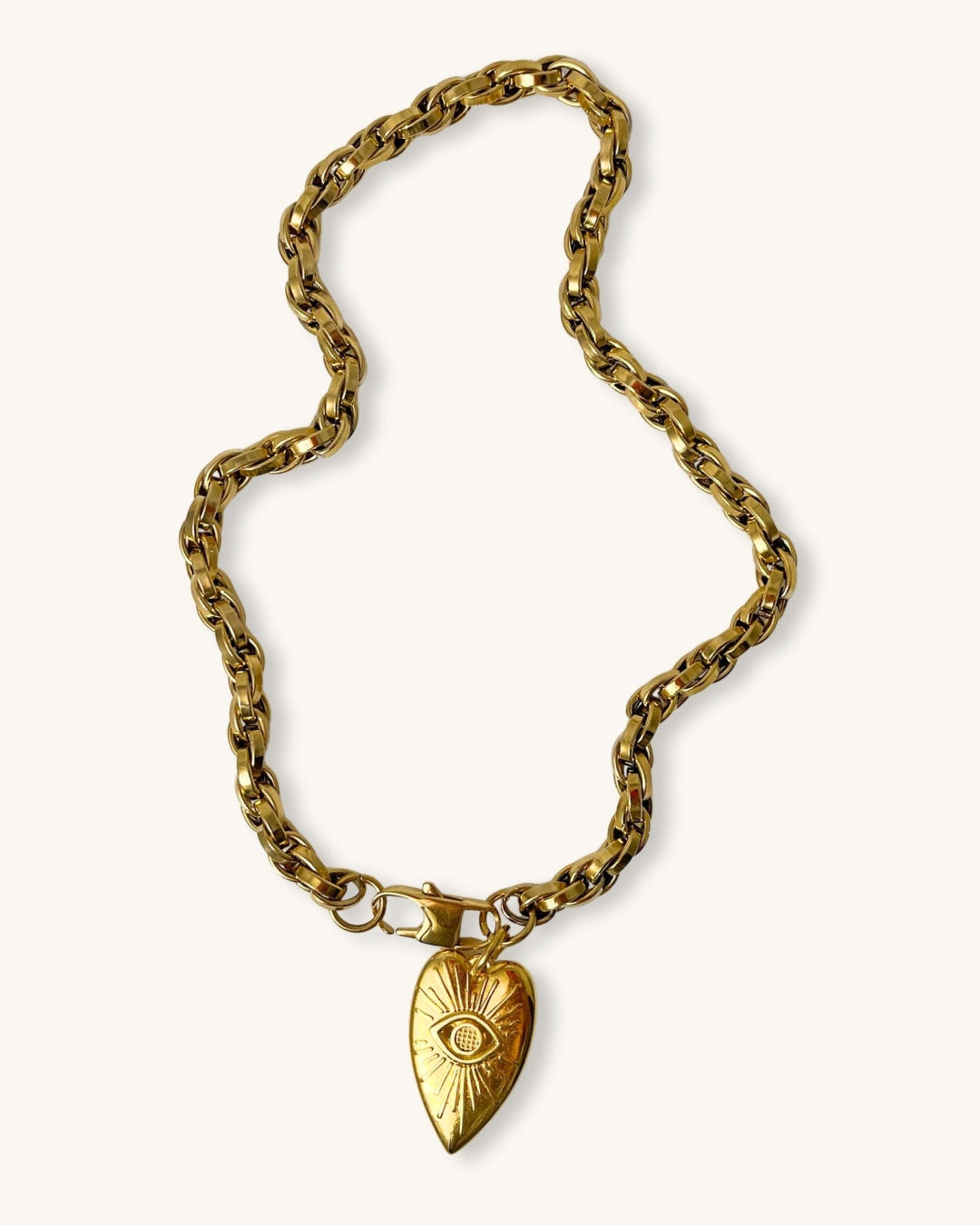 A gold chain necklace with a silver heart pendant against a neutral background. The heart pendant has a raised eye design with sun rays around the eye.