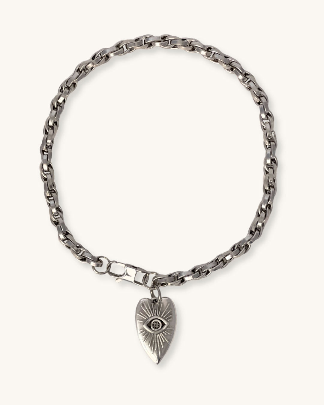 A silver chain necklace with a silver heart pendant against a neutral background. The heart pendant has a raised eye design with sun rays around the eye.