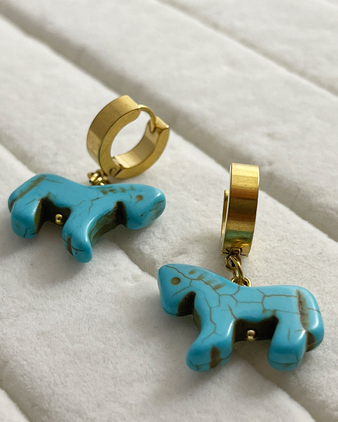 Turquoise horse-shaped earrings with gold hoops on a white fabric background