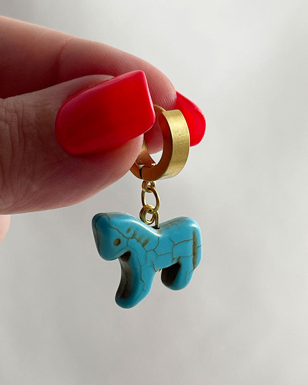 Gold hoop earring with a turquoise horse charm held by a hand against a white background