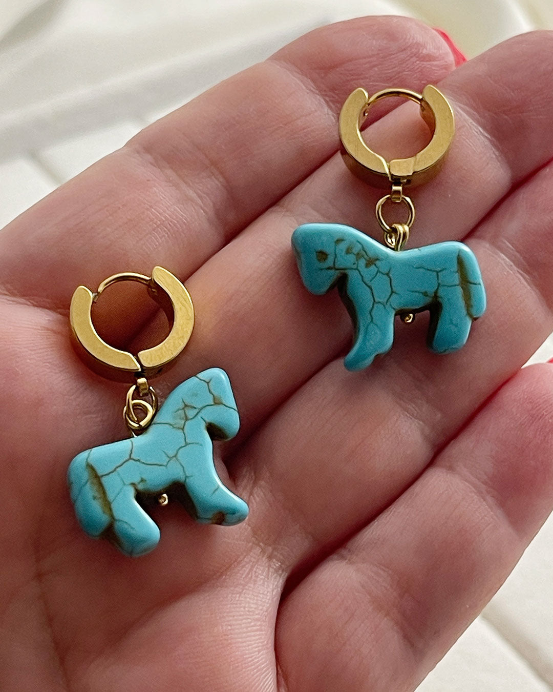 Turquoise horse-shaped earrings with gold hoops on a hand.