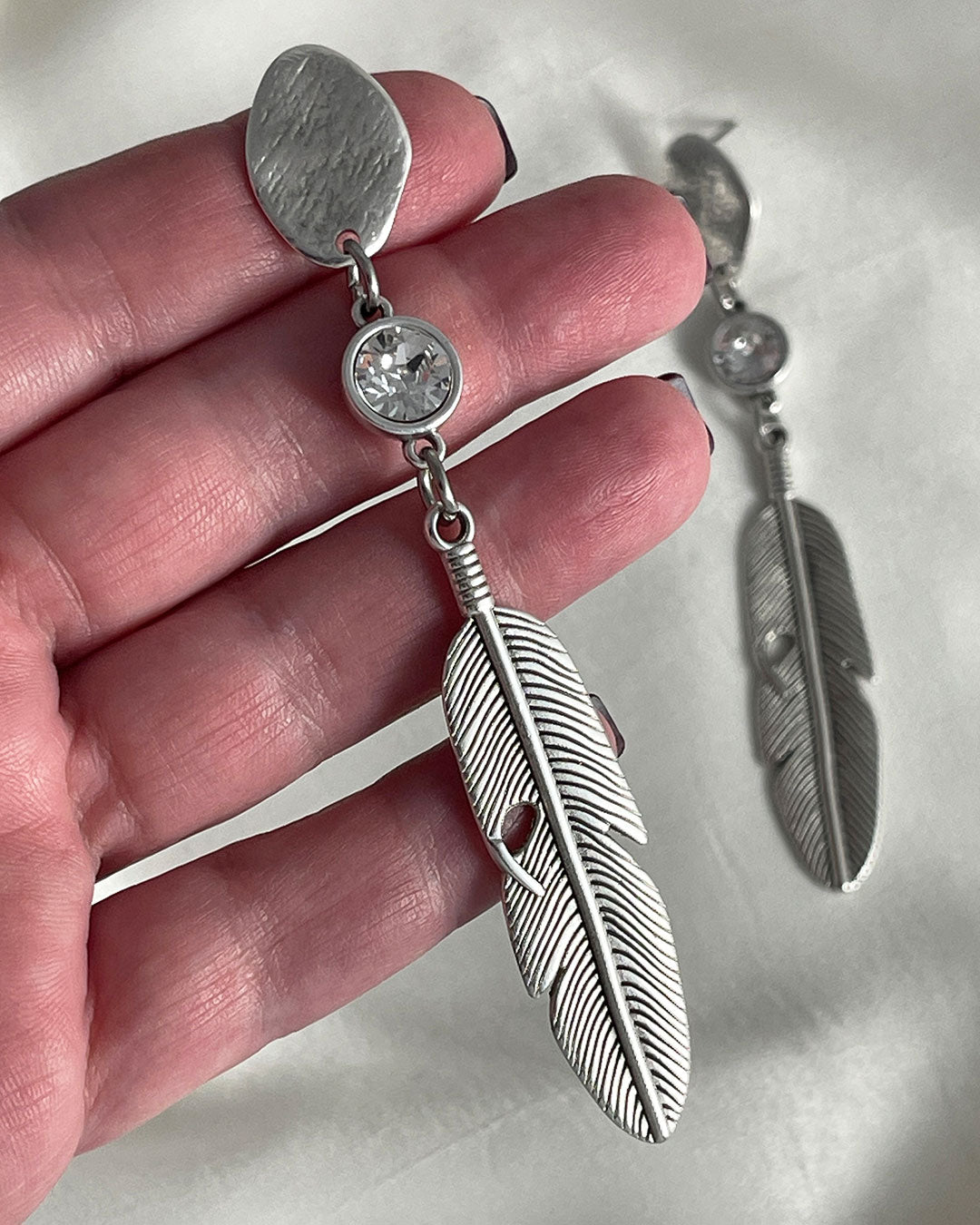 A pair of silver feather earrings with a crystal clear Swarovski. Each earring features a textured silver feather charm dangling from silver oval-shaped studs. The earrings have a bohemian style and are suitable for adding a touch of elegance to any outfit.