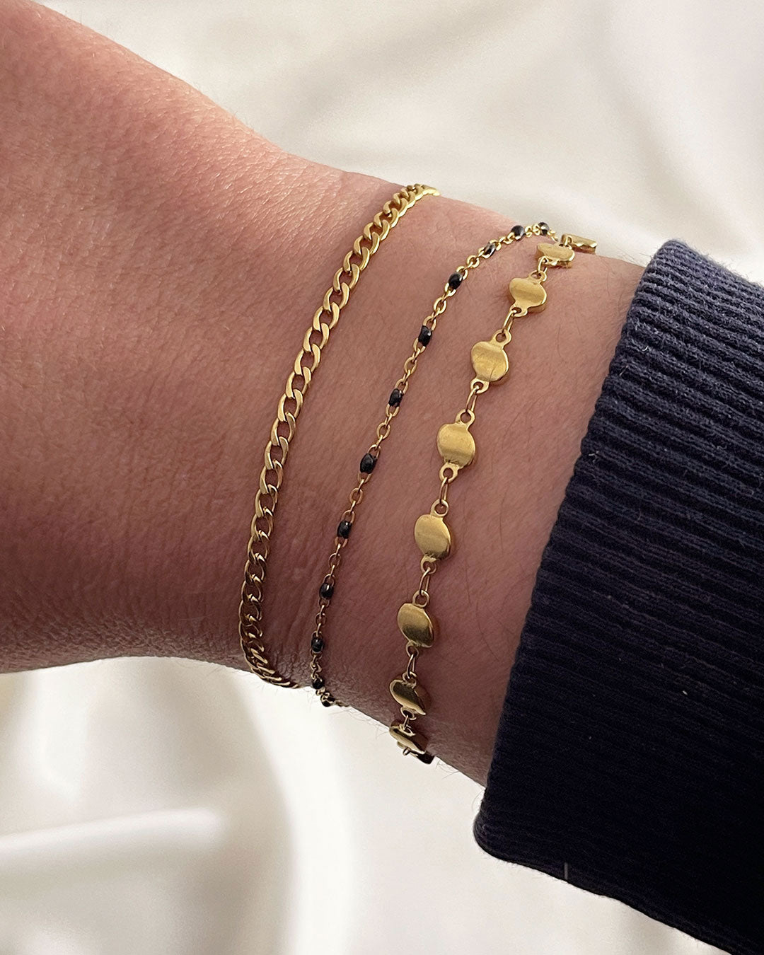 Model wearing three gold bracelets stacked, a curb chain bracelet, a beaded black enamel bracelet, and a flat beaded chain bracelet.