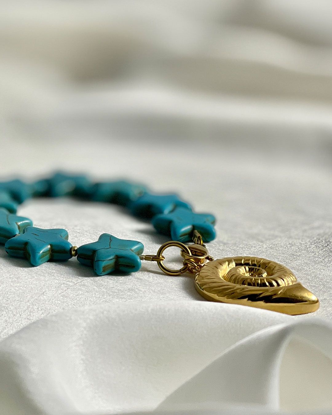 Bracelet with turquoise star-shaped howlite beads and a large gold nautilus shell charm on a white fabric background