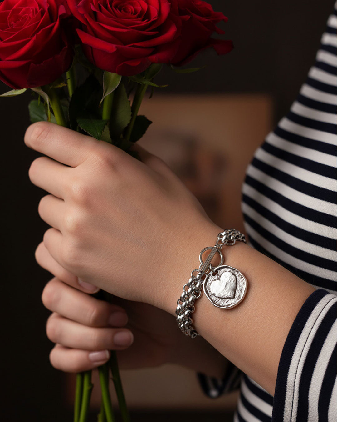 Hand holding a bouquet of red roses with a silver chainmail bracelet featuring a heart charm.