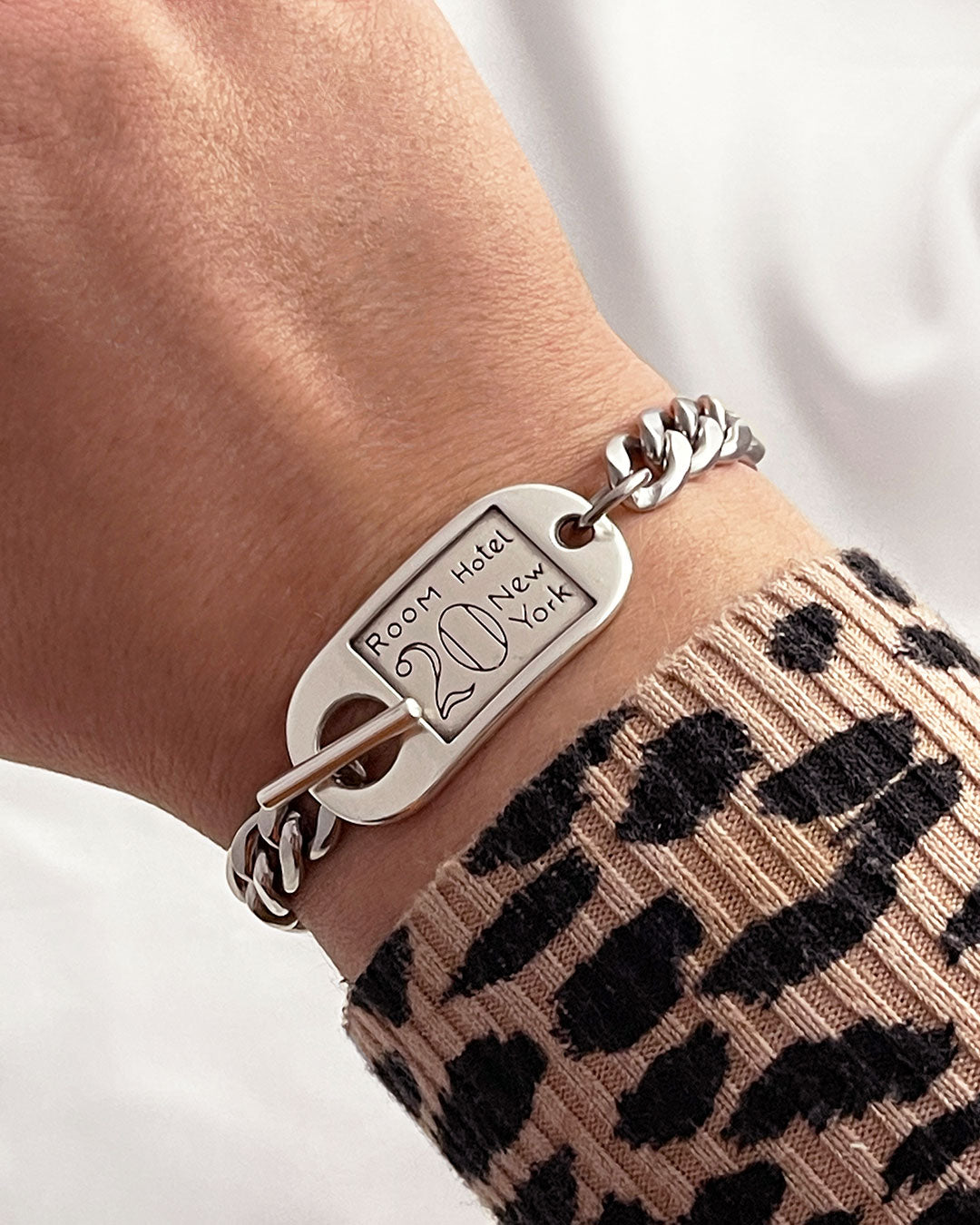 Model showcases a silver cuban curb chain bracelet with a silver hotel key tag charm. The key tag has the engraved text: Room Hotel, 20, New York.