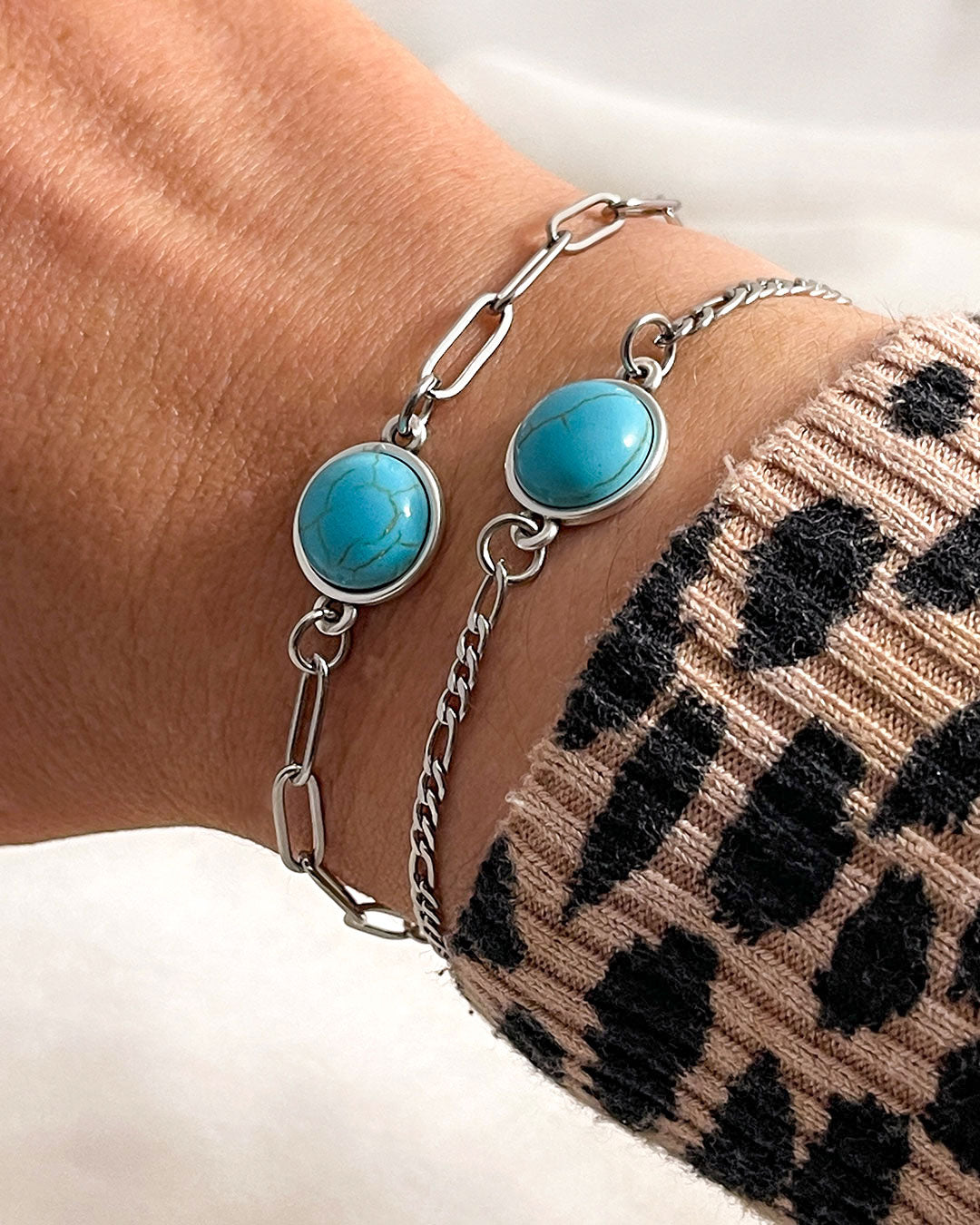 Two silver bracelets with turquoise stones, one with a paperclip and the other with a figaro chain on a wrist against a neutral background