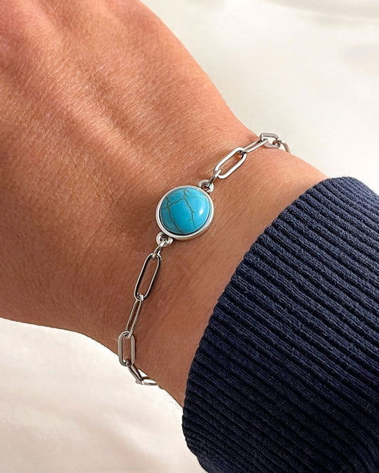 Turquoise stone bracelet with paperclip chain in silver on a wrist with a black sleeve against a white background