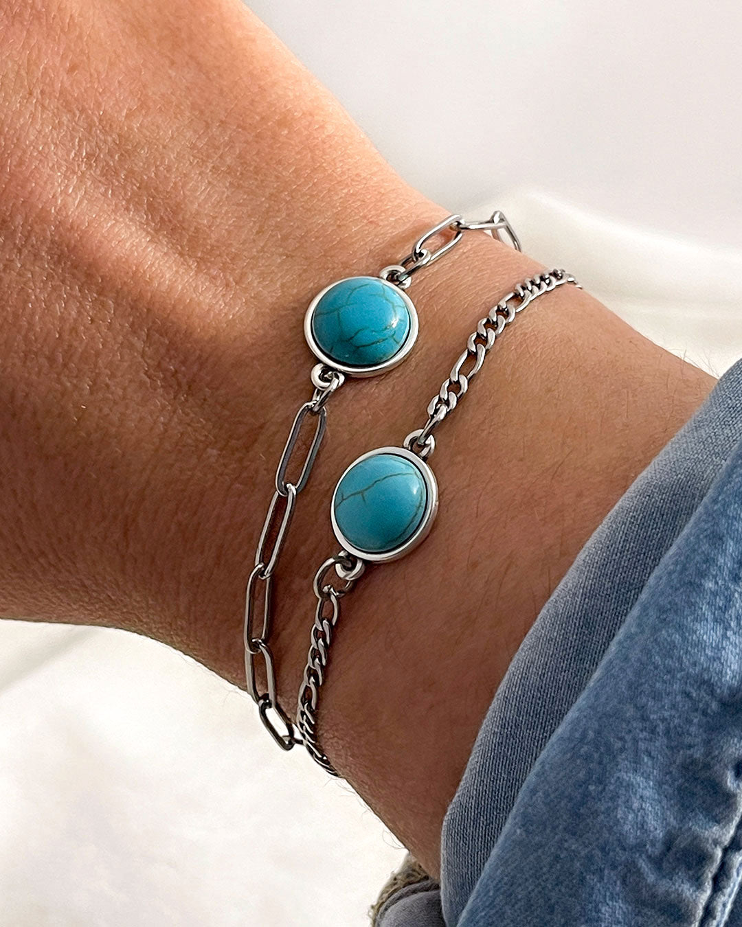 Two silver bracelets with turquoise stones, one with a paperclip and the other with a figaro chain on a wrist against a neutral background