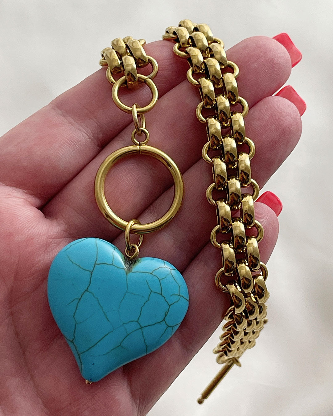 Chunky gold chain with a turquoise heart pendant held in a hand against a neutral background