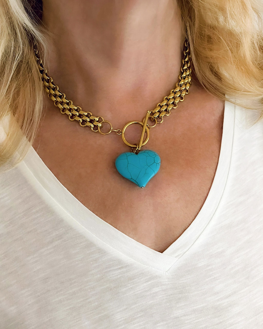 Chunky gold chain necklace with a turquoise heart pendant worn by a person in a white shirt.