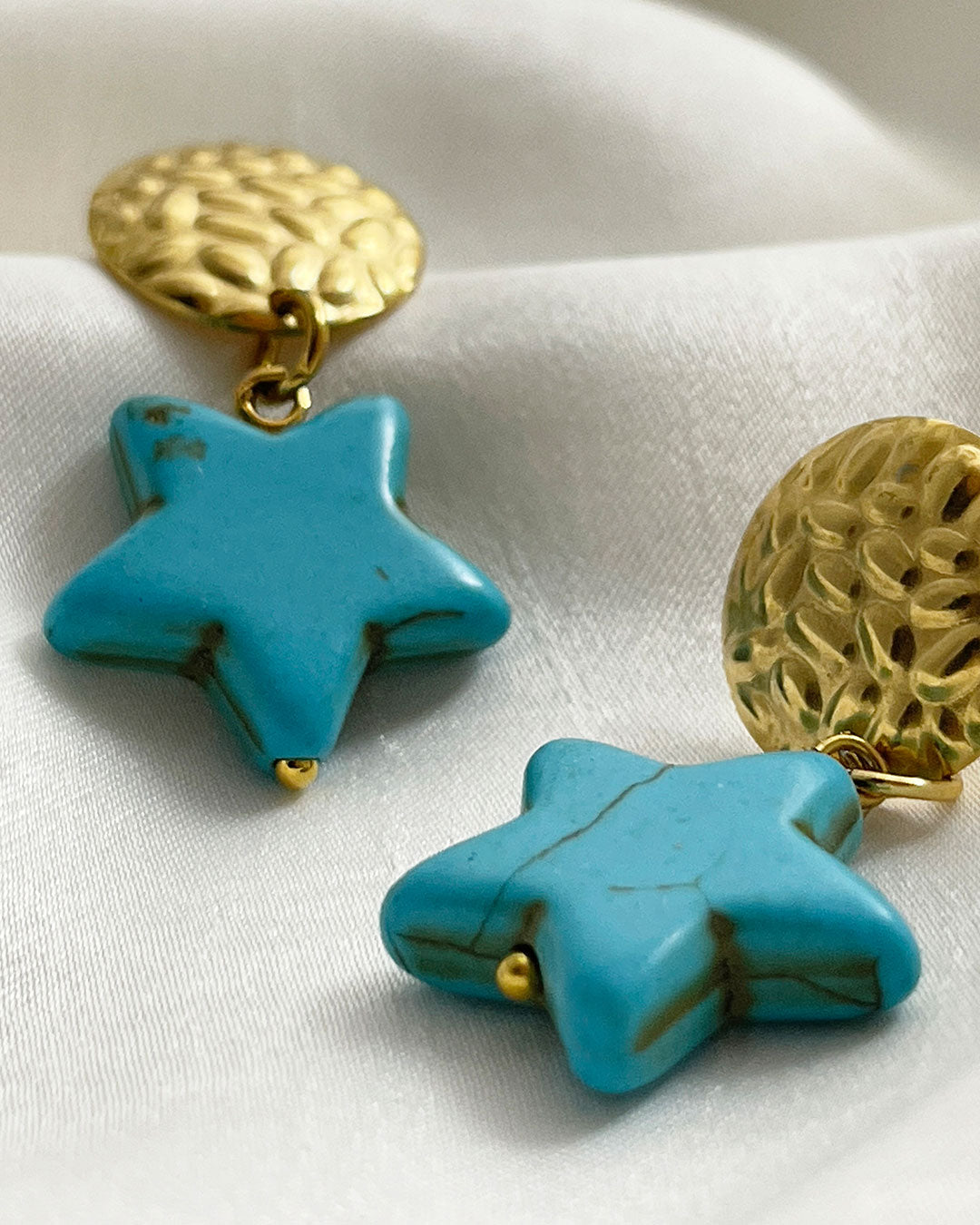 Pair of drop earrings with turquoise star pendants and gold circular studs on a white fabric background.