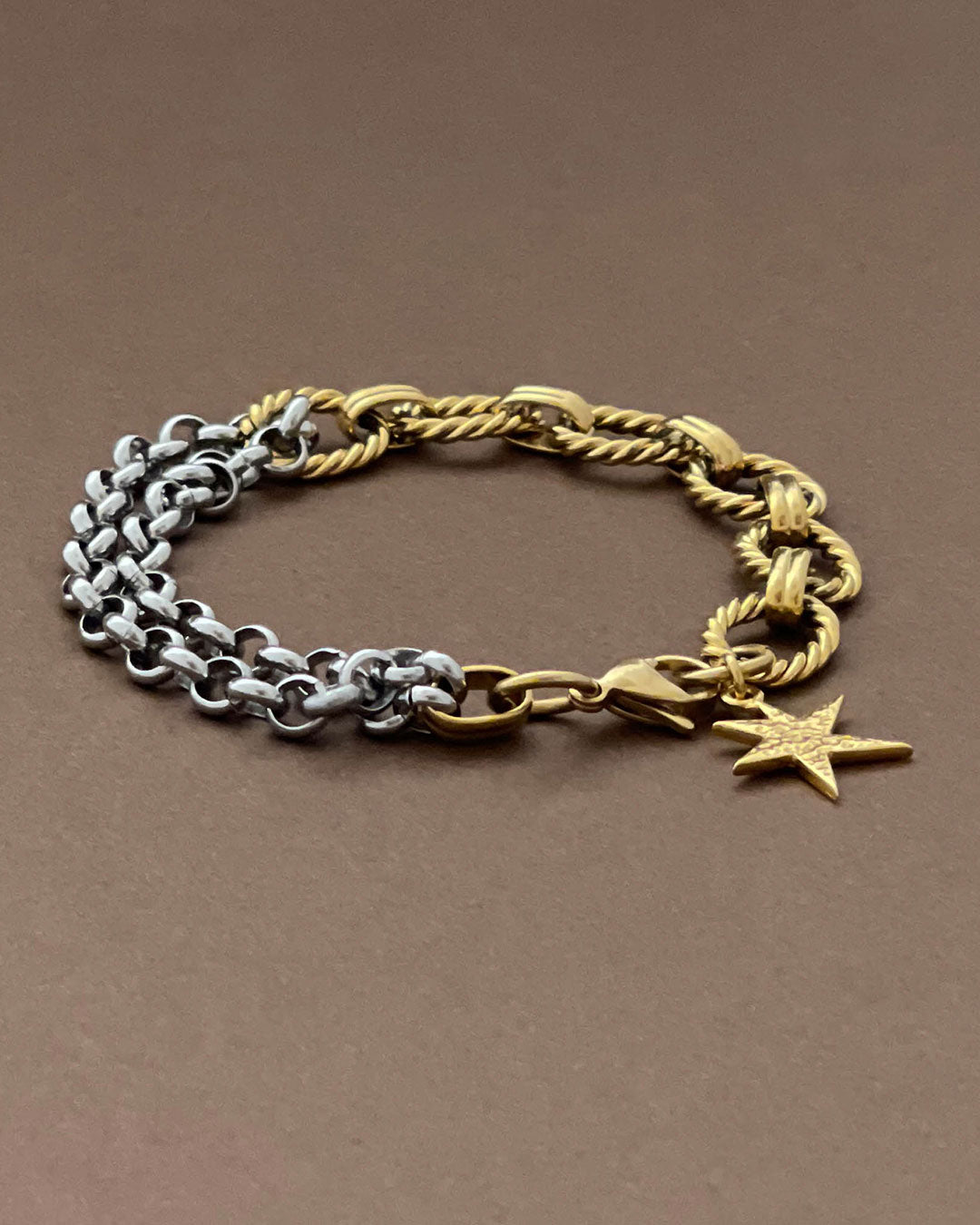 Two chain bracelets, one gold and one silver with a gold charm charm on a brown background