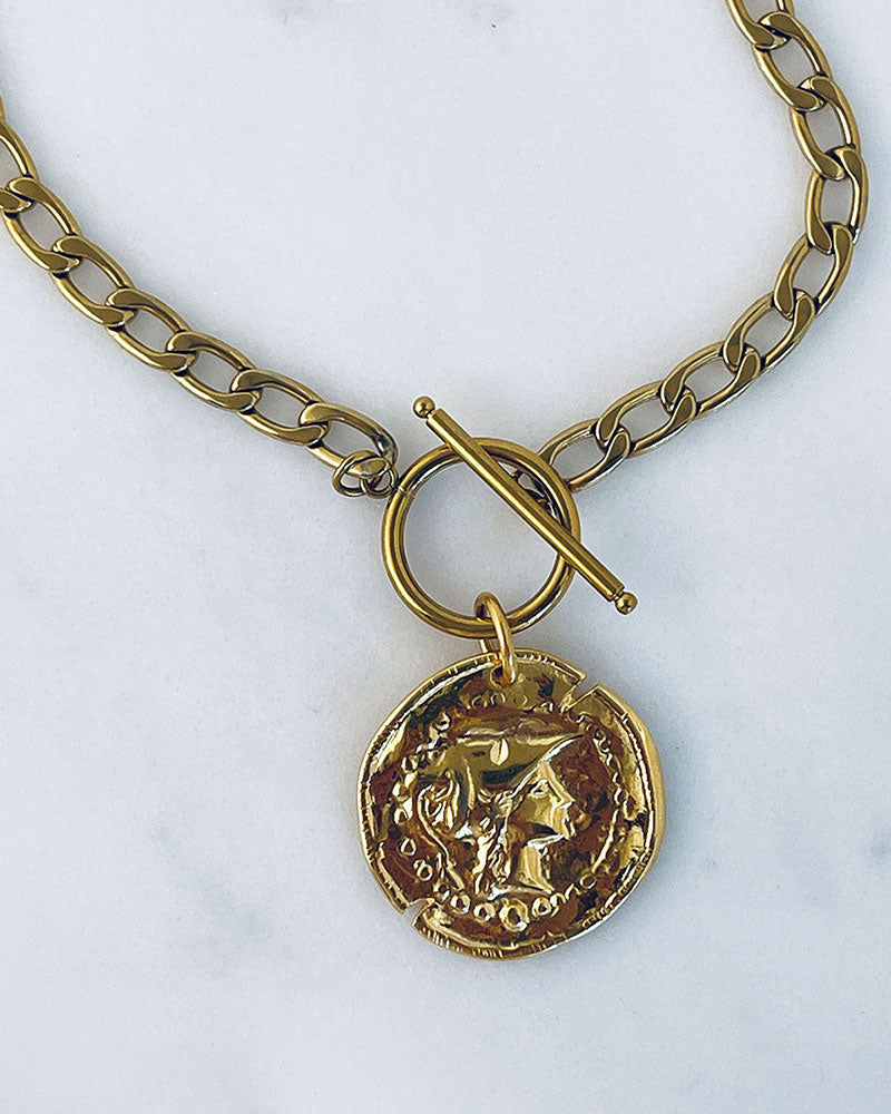 A gold chain necklace with a large, round, gold coin pendant. The coin has a raised design of Athena's goddess face. The chain is a curb chain style with a toggle clasp.