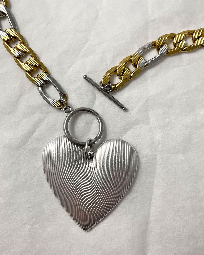 A bold statement chain necklace with a large textured silver heart pendant. The two-tone stainless steel figaro chain blends silver and gold for a striking contrast of luxury.