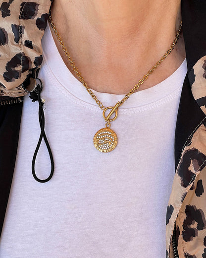 Model wearing a gold chain necklace with a round pendant featuring a design of a Horus eye with small clear rhinestones. The pendant is attached to the chain with a toggle clasp.