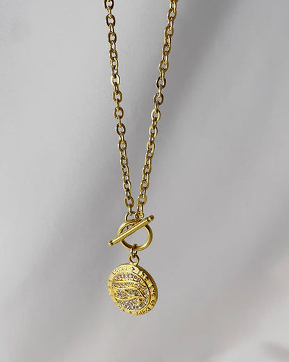 A gold chain necklace with a round pendant featuring a design of a Horus eye with small clear rhinestones. The pendant is attached to the chain with a toggle clasp.