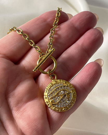 A gold chain necklace with a round pendant featuring a design of a Horus eye with small clear rhinestones. The pendant is attached to the chain with a toggle clasp.