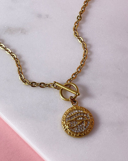 A gold chain necklace with a round pendant featuring a design of a Horus eye with small clear rhinestones. The pendant is attached to the chain with a toggle clasp.