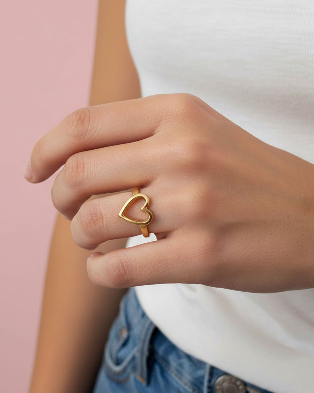 Gold heart-shaped ring on a person's finger against a pink background