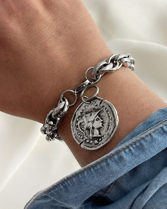 Chunky silver bracelet with an Athena coin charm on a wrist, against a neutral background