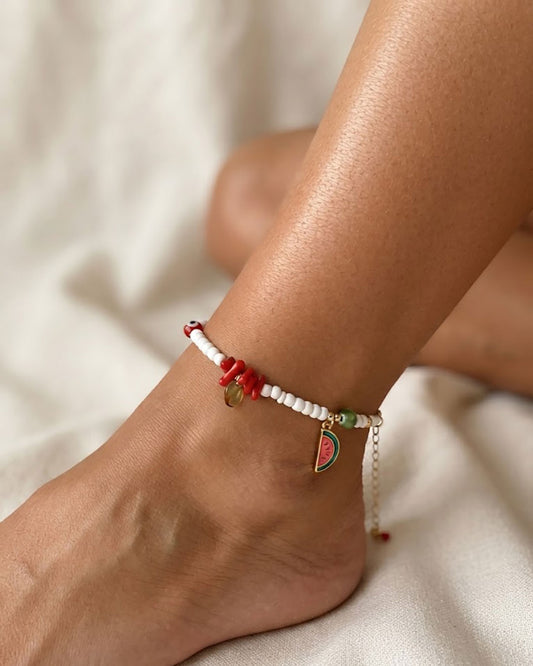 A close-up of a woman's ankle and foot wearing a delicate beaded anklet with red and white beads, red corals, and a watermelon slice charm, against a soft beige fabric background.