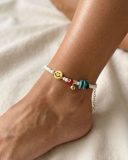 A white beaded anklet adorned with a yellow smiley happy face bead and turquoise shell chips on a woman ankle