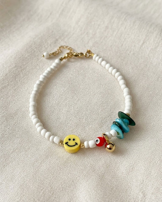 A white beaded anklet adorned with a yellow smiley happy face bead and turquoise shell chips on a fabric backround