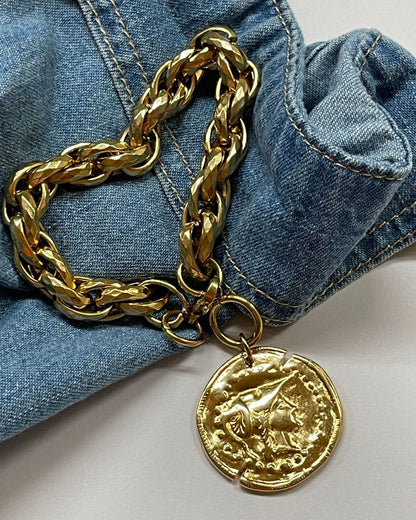 A gold chain bracelet with a large, round, gold coin charm. The coin has a raised design of Athena's goddess face. 
