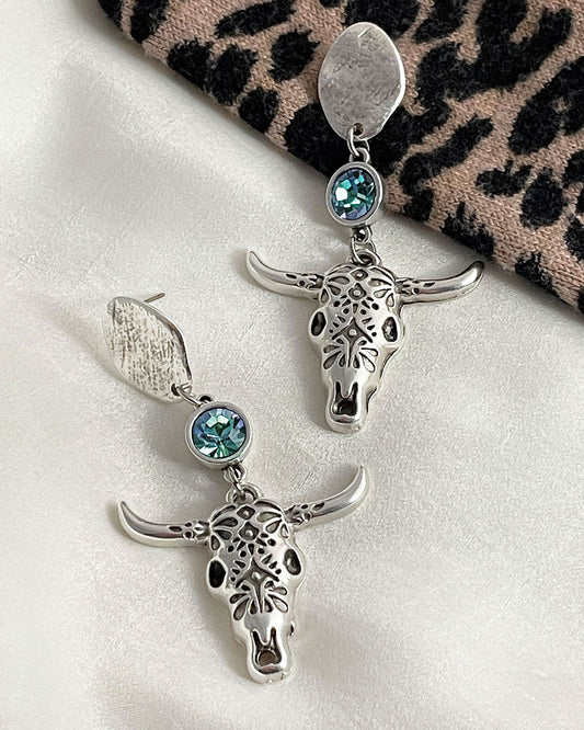 A pair of silver earrings featuring longhorn bull skull large charms. Each pendant is detailed with an embossed design and accented with an aquamarine Swarovski crystal. The earrings hang from silver oval-shaped studs. These Western-style earrings are perfect for adding a touch of bohemian flair to any outfit.