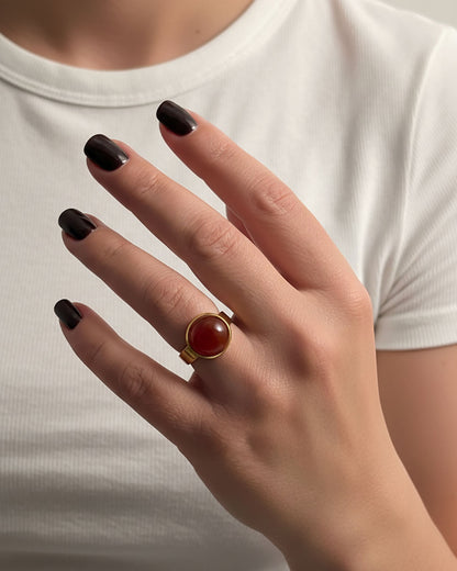 Hand wearing a gold ring with a carnelian gemstone against a neutral background