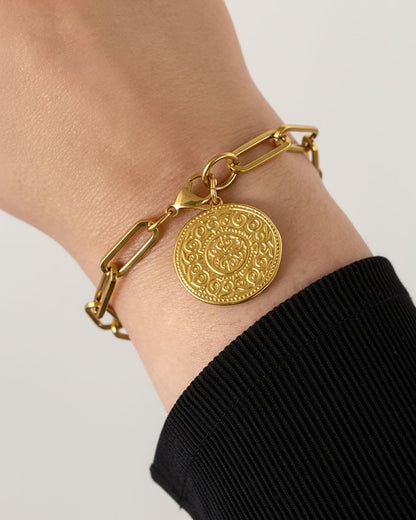 Gold bracelet with a decorative coin charm on a wrist against a neutral background