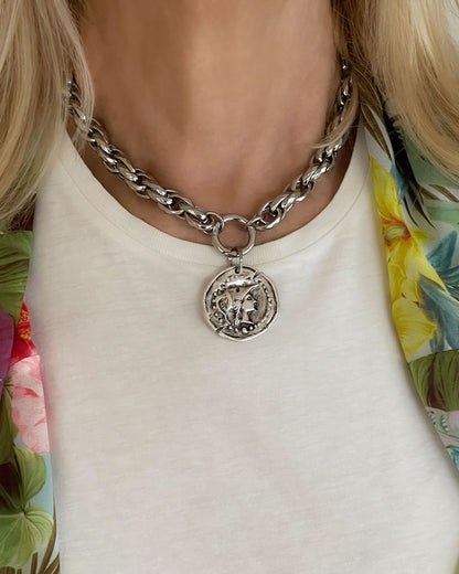 Model wearing a silver tone chunky chain necklace with a large coin pendant. The coin features an embossed profile of a woman's face.  The necklace is a bold statement piece with a substantial chain and a detailed pendant. The chain is thick and rope-like, and the pendant is substantial and eye-catching.