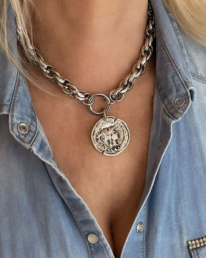 Model wearing a silver tone chunky chain necklace with a large coin pendant. The coin features an embossed profile of a woman's face.  The necklace is a bold statement piece with a substantial chain and a detailed pendant. The chain is thick and rope-like, and the pendant is substantial and eye-catching.
