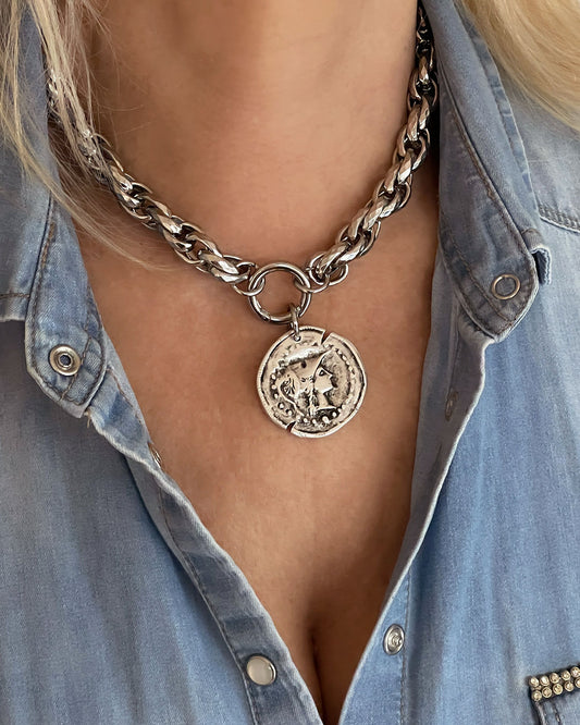 Model wearing a silver tone chunky chain necklace with a large coin pendant. The coin features an embossed profile of a woman's face.  The necklace is a bold statement piece with a substantial chain and a detailed pendant. The chain is thick and rope-like, and the pendant is substantial and eye-catching.