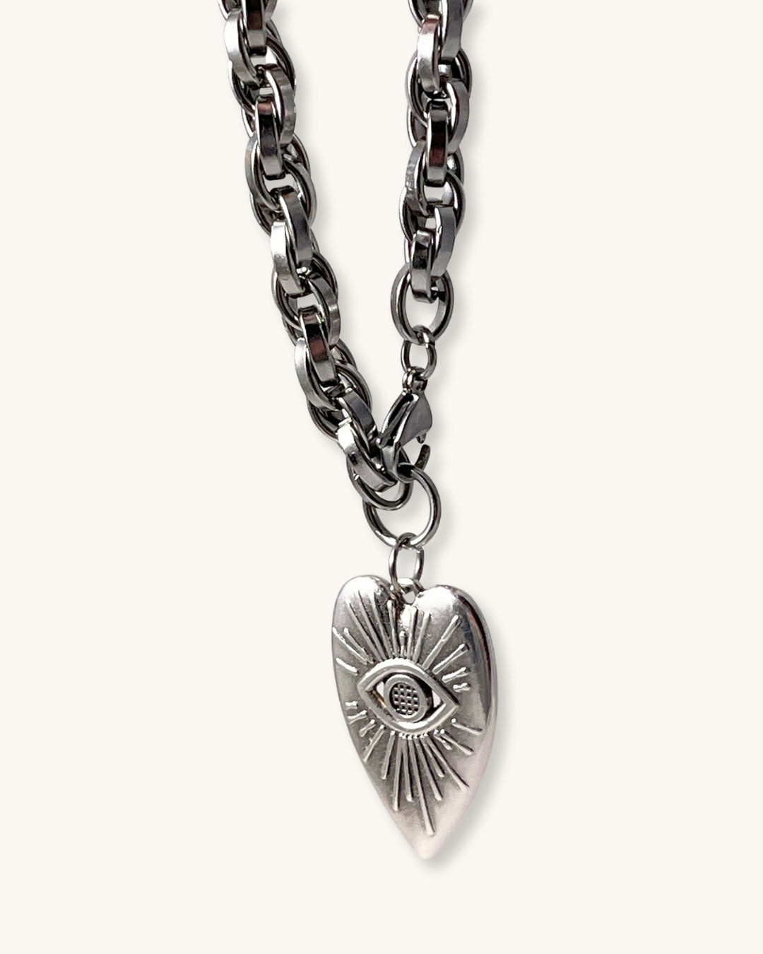 A silver chain necklace with a silver heart pendant against a neutral background. The heart pendant has a raised eye design with sun rays around the eye.