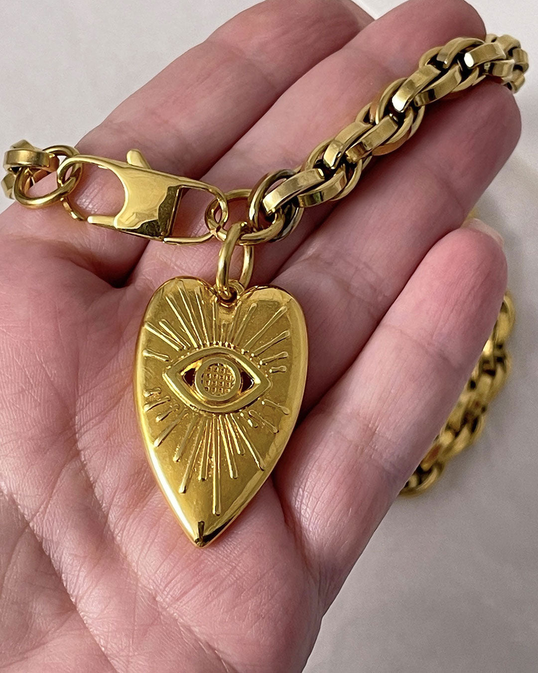 A hand holds a gold chain necklace with a silver heart pendant against a neutral background. The heart pendant has a raised eye design with sun rays around the eye.